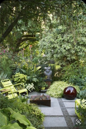 A modern tranquil shady garden with concrete and gravel "flooring", pale yellow/green furniture, and foliage that is mostly greens and yellows.